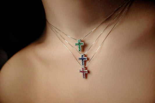 La foto mostra una modella che indossa 3 collane con 3 croci. UN croce mota smeraldi e diamanti, una monta zaffiri e diamanti e la terza rubini e diamanti. 