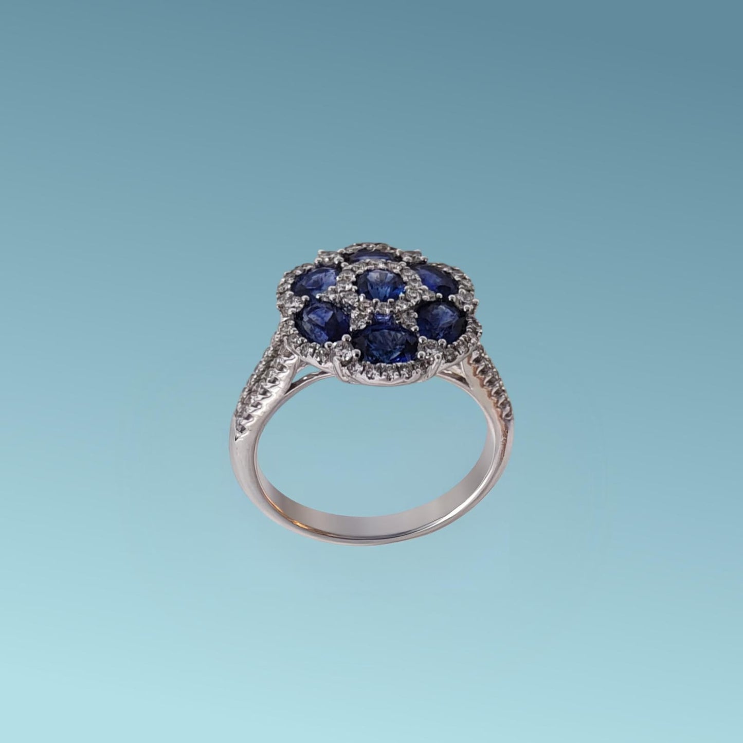 anello con uno zaffiro blu naturale dal taglio brillante incastonato al centro, circondato da sei zaffiri blu naturali dal taglio ovale per un totale di 2.18 ct, tutti abbracciati da un pavé di diamanti naturali taglio brillante g/vs